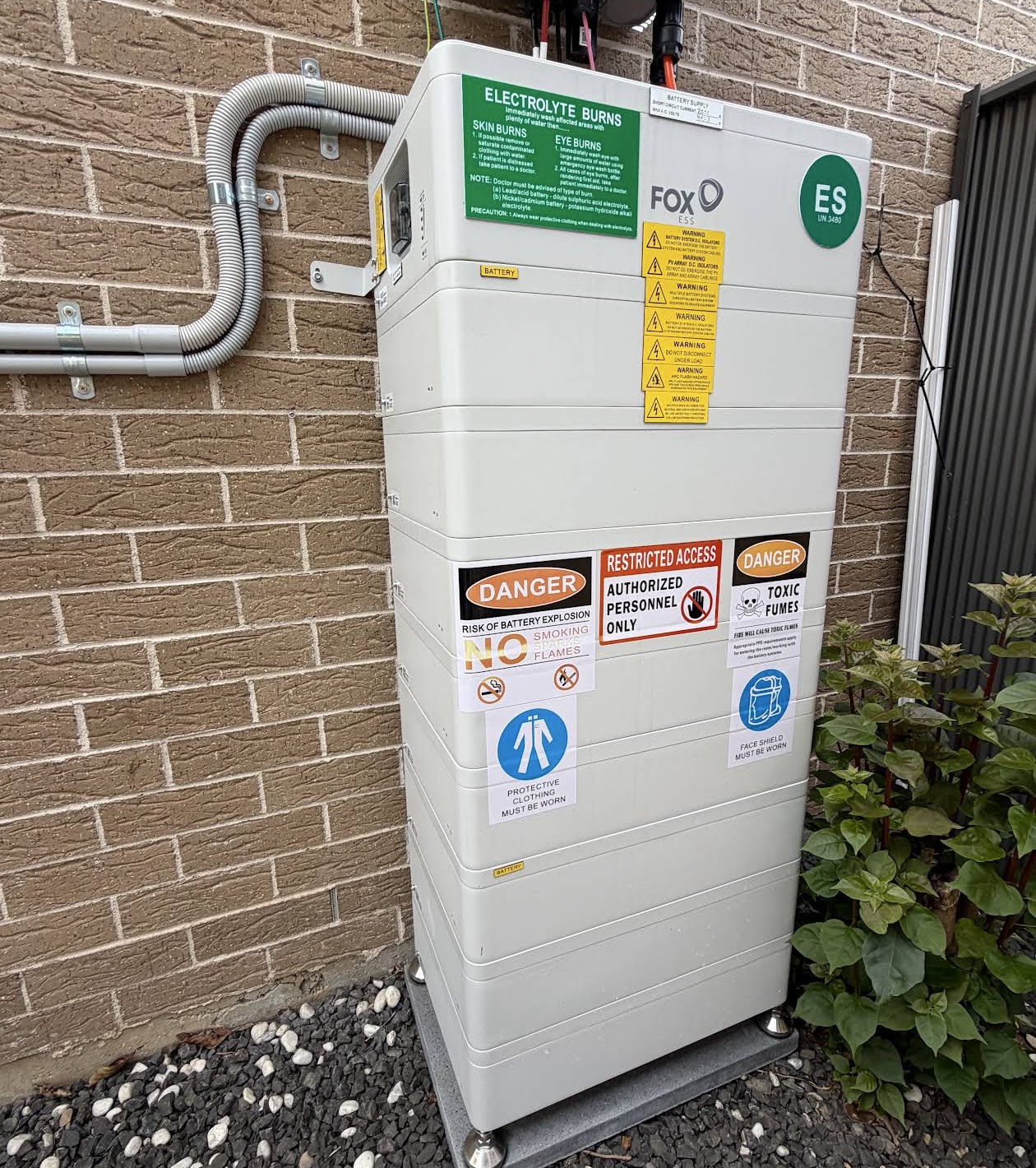 Home battery storage unit installed against a brick wall with conduit and safety signage
