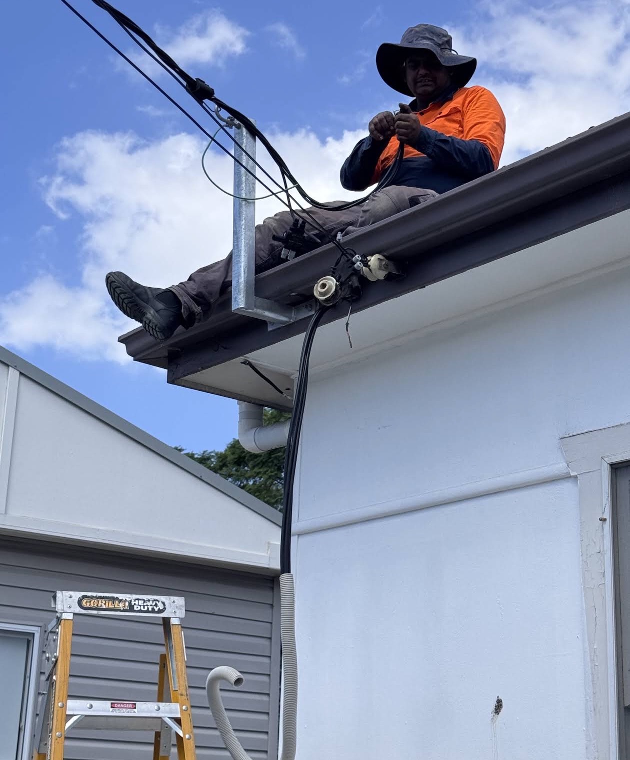 Electrician in hi-vis sitting on a house roofline handling the overhead service cable, ladder below